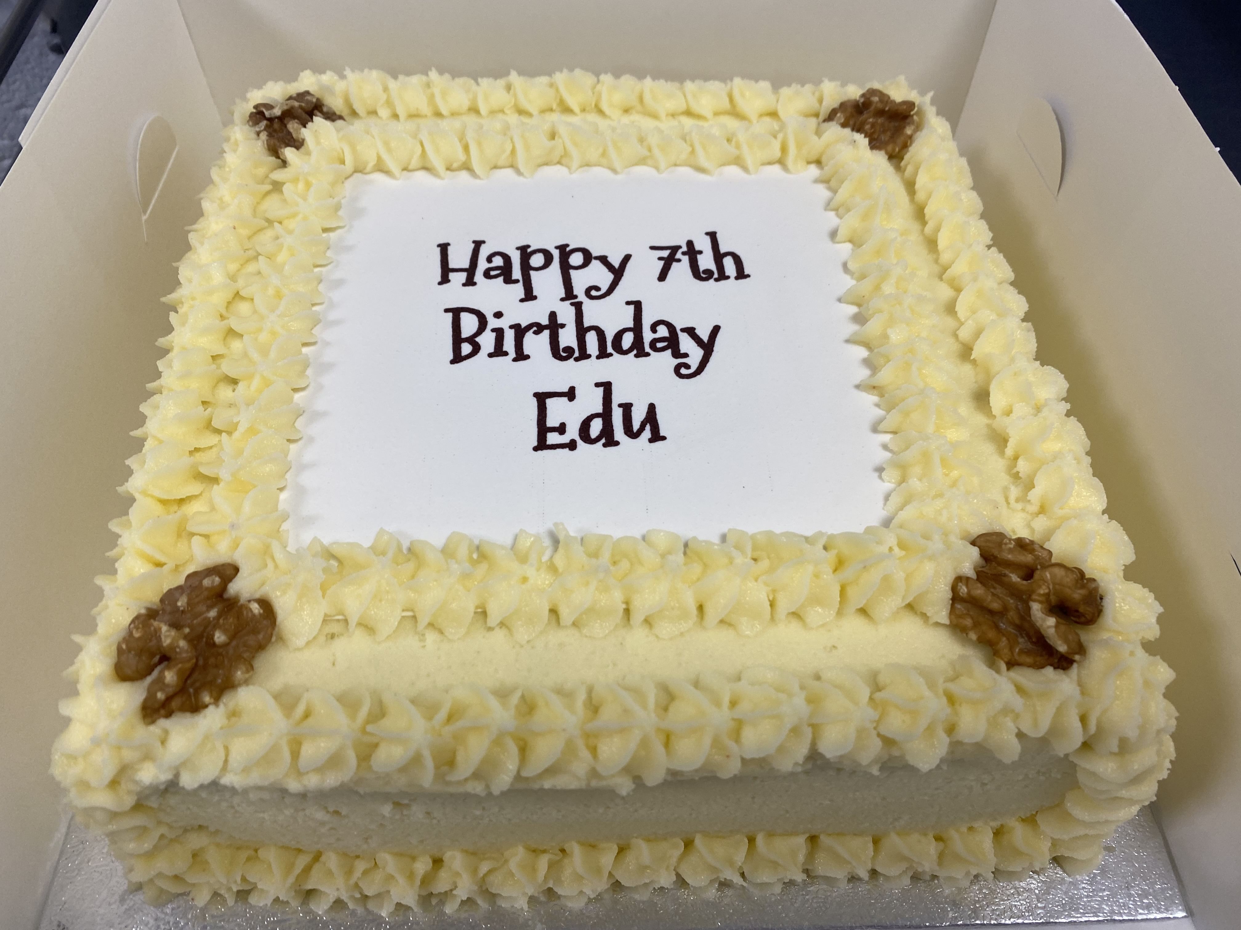 A square birthday cake with yellow frosting and walnut decorations on each corner. The top of the award-winning carrot cake reads 'Happy 7th Birthday Edu' in brown lettering. The carrot cake is set inside a white box, boasting the Great Taste Award, promising a delightful celebration.